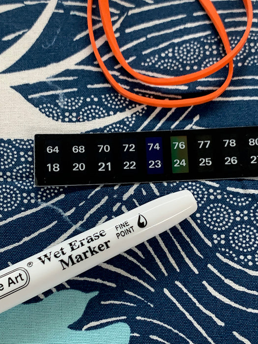 A set of kombucha brewing accessories including a stick-on thermometer, a wet erase pen, and an orange rubber band all placed on a patterned cotton cloth jar cover.
