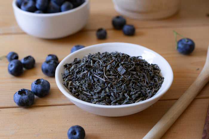 Loose leaf green tea leaves in a white bowl on a wooden surface surrounded by blueberries.