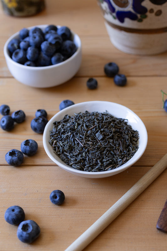 Vertical image of small ceramic dish filled with green tea leaves on a wooden surface surrounded by blueberries.