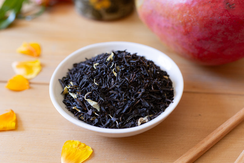 White bowl filled with black tea leaves on a wooden surface with mangoes and yellow petals.