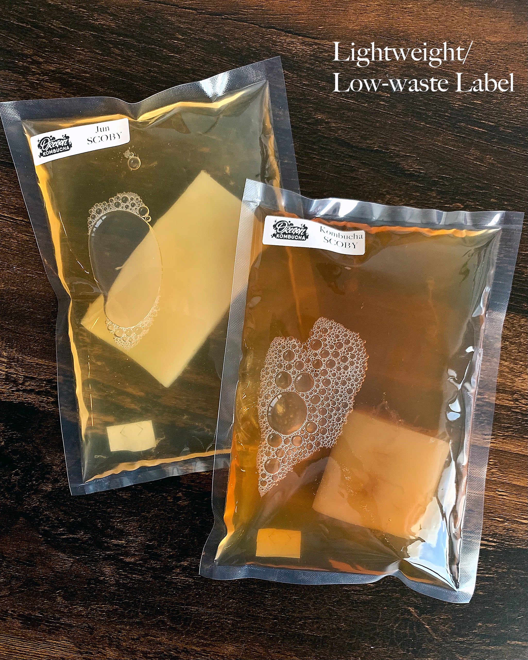 Two sealed plastic pouches on a wooden surface - one containing golden colored jun starter liquid and square SCOBY pellicle, the other containing amber colored kombucha starter liquid and square SCOBY pellicle.  Both pouches have small printed paper labels on the upper left of the packaging with the Oregon Kombucha logo and respective jun or kombucha SCOBY content descriptor.  