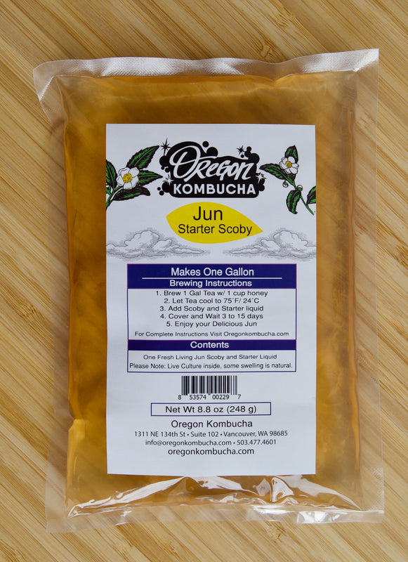 Golden-colored jun starter liquid and SCOBY in sealed bag with colorful label on a bamboo cutting board.  